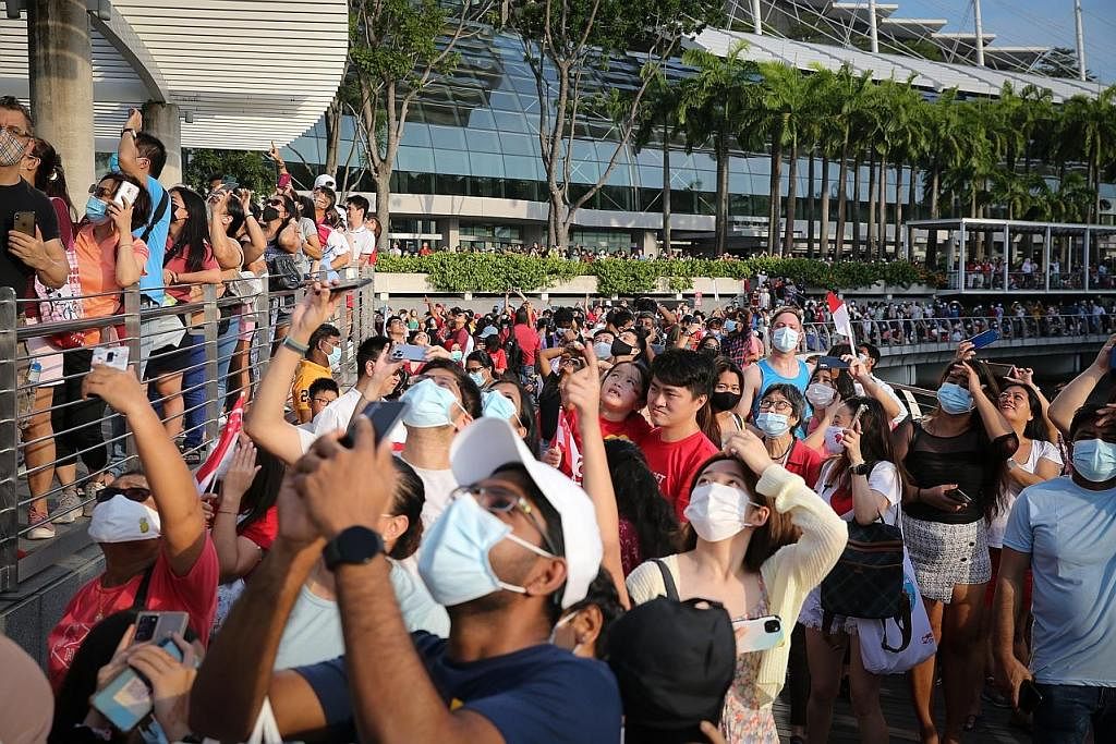 MENYANYI LAGU KEBANGSAAN: Penonton berkumpul sambil memegang bendera Singapura di luar Marina Bay Sands untuk menyaksikan acara Terbang Lalu Bendera Negara. - Foto-foto BH oleh ONG WEE JIN SAMA-SAMA RAIKAN KEMERIAHAN: Pasangan dan kumpulan penduduk i
